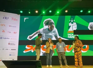 Carla Fonseca, da GOL, no palco do evento de lançamento da Taça das Favelas São Paulo 2024, com os mestres de cerimônia Hélio de la Peña (à esq.) e Letícia Vidica (à dir.) (Foto: Divulgação GOL)
